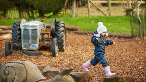 A child dressed for winter in pink wellies and a bobble hat jumping across log stepping stones